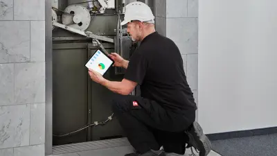 A technician kneeling in front of an elevator with tablet computer in hand uses the Bosch Elevator Cloud to analyze the elevator's data